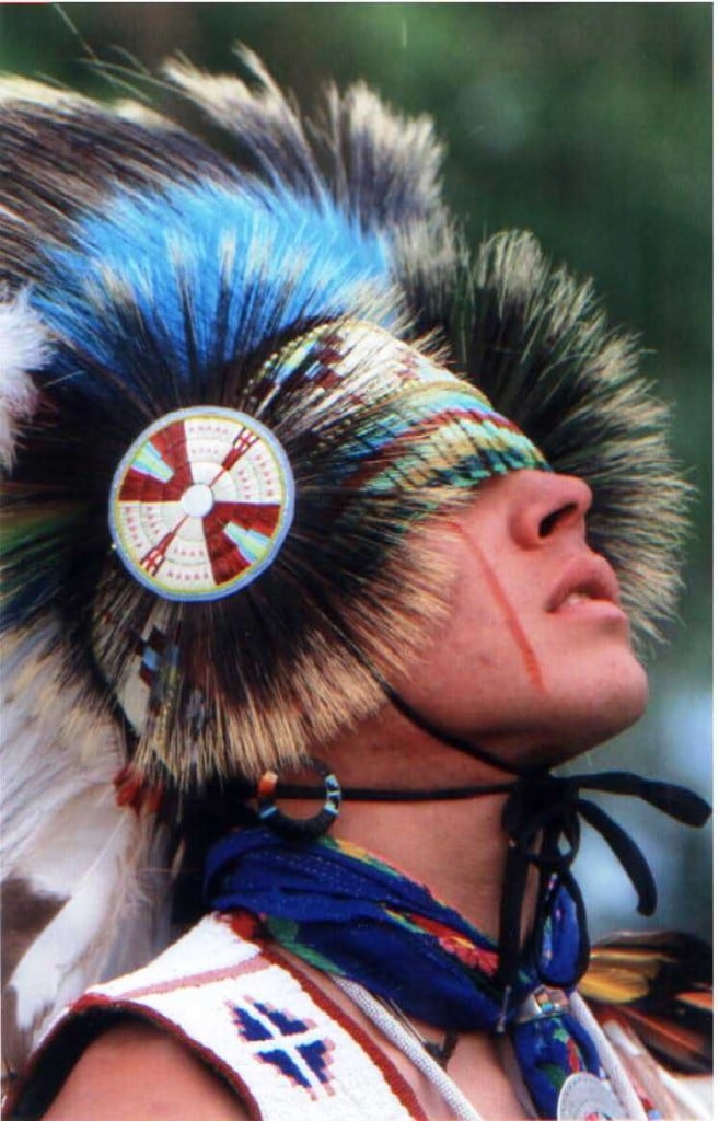 A person wearing a colorful feathered headdress and traditional attire looks upward, with face paint and beaded accessories visible.