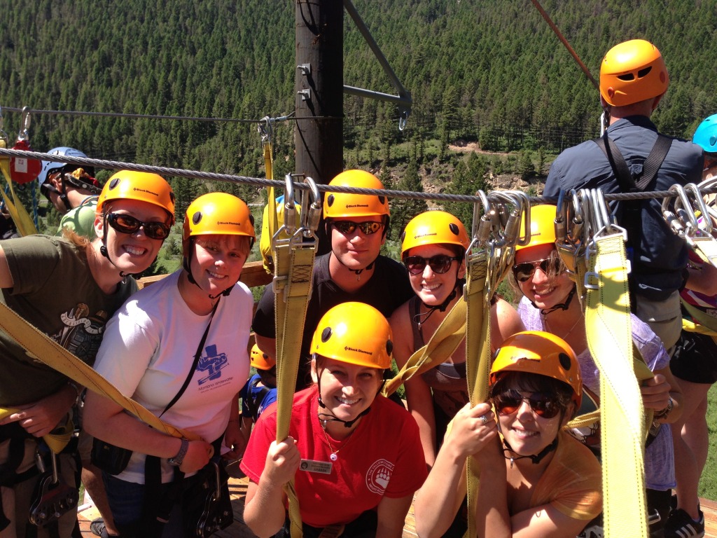 A group of people, including a Yellowstone employee, wearing yellow helmets and harnesses stand together outdoors, preparing for an outdoor recreation ziplining activity in a forested area.