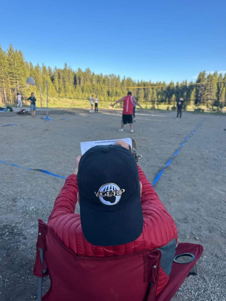 A Canyon employee at the Yellowstone recreation center, wearing a red jacket and black hat, sits in a chair writing on a clipboard while observing a volleyball game on a dirt court with trees in the background.