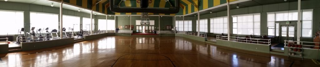 Panoramic view of an empty indoor gymnasium with a basketball court, exercise machines on the left, benches on the right, and large windows along the walls.