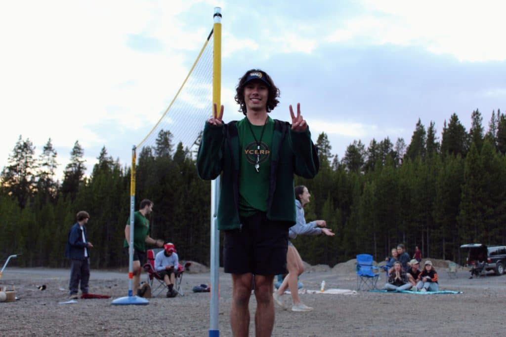 A young Yellowstone employee stands by a volleyball net outdoors, making peace signs with both hands. Several others gather in the background on a gravel area near trees, enjoying casual athletics together.