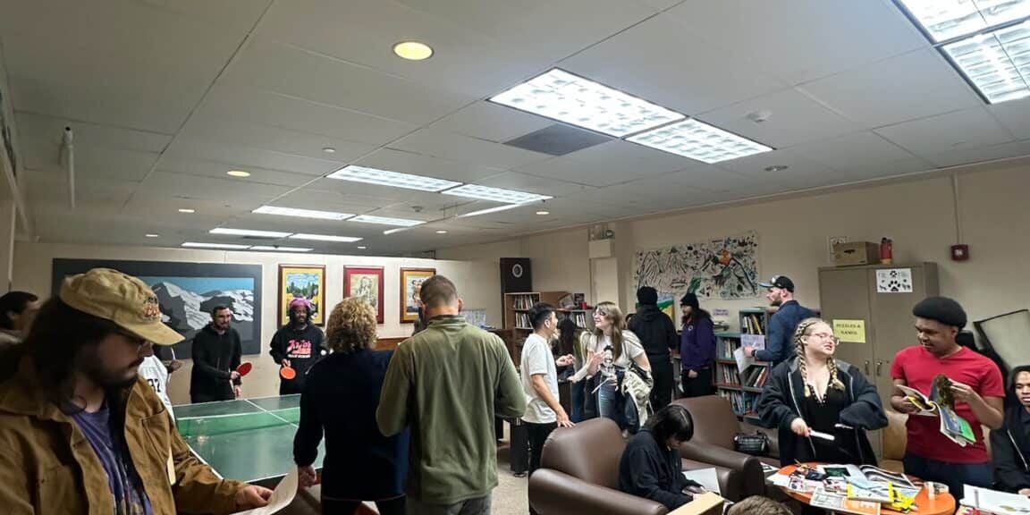 A group of people socialize in a lounge with a ping pong table, couches, snacks, and games visible on tables.