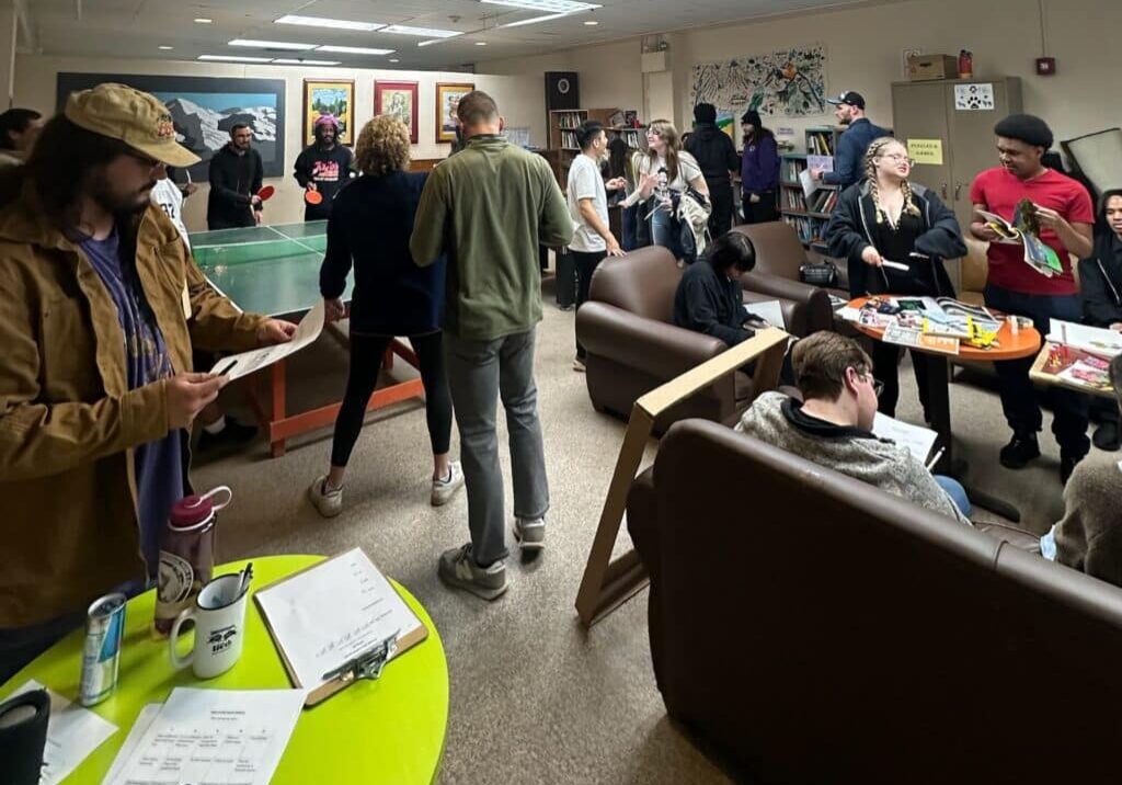 A group of people socialize in a lounge with a ping pong table, couches, snacks, and games visible on tables.