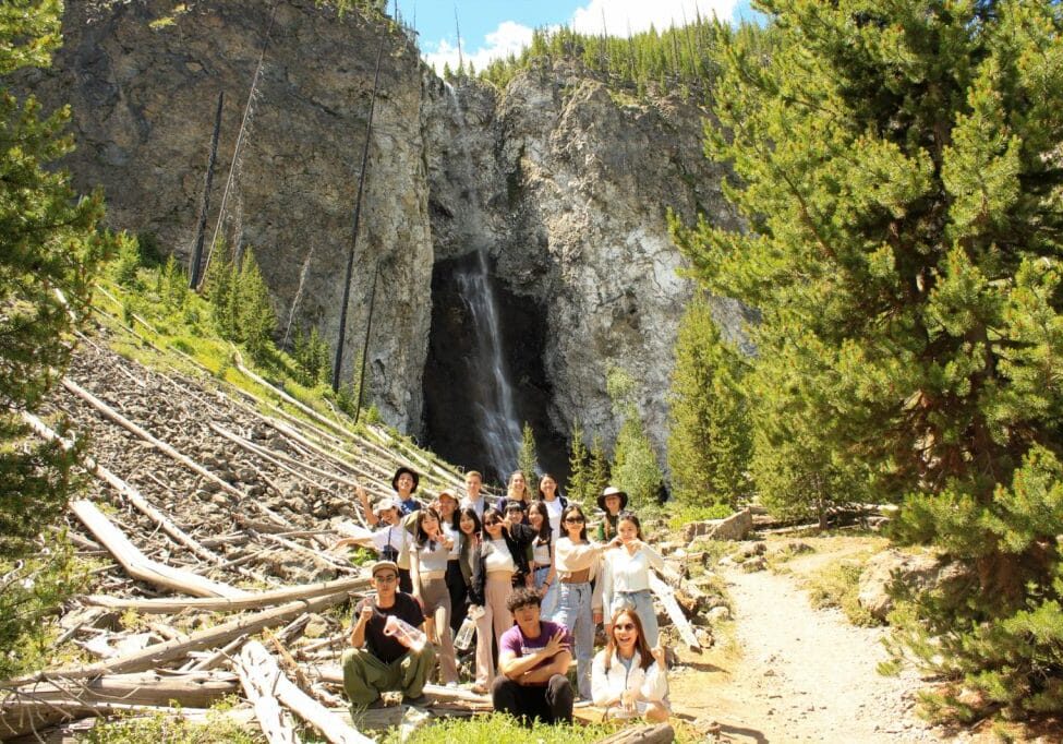 A group of people pose for a photo on a trail with a waterfall and rocky cliffs in the background, surrounded by trees and fallen logs.