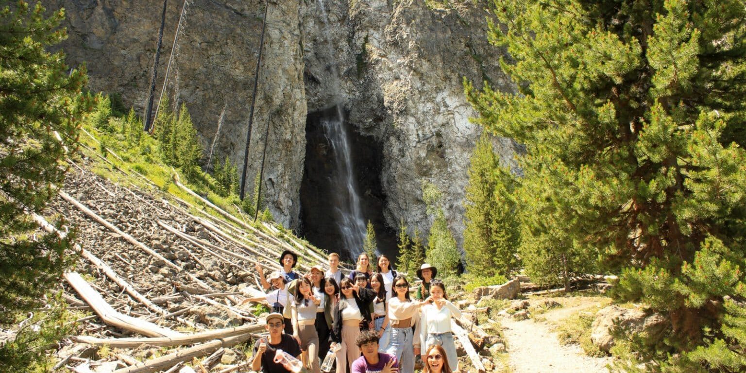 A group of people pose for a photo on a trail with a waterfall and rocky cliffs in the background, surrounded by trees and fallen logs.