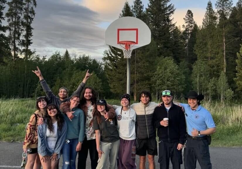 A group of eleven people stand together smiling under a basketball hoop outdoors, with trees and grass in the background.