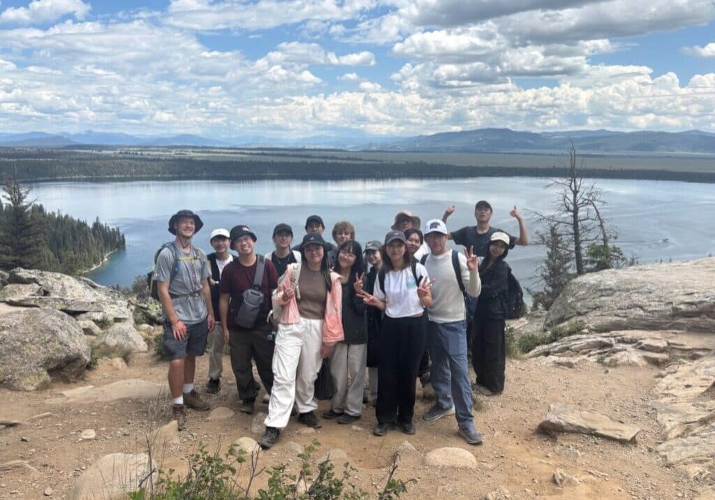 A group of Yellowstone employee hiking club members stands on a rocky overlook with a lake and forested landscape in the background under a partly cloudy sky.