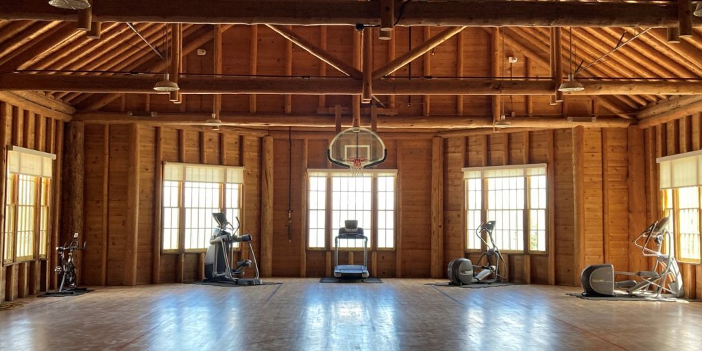 Wood-paneled gym at the Lake employee recreation center in Yellowstone, featuring a basketball hoop, treadmill, and various exercise machines arranged under exposed beams and large windows.