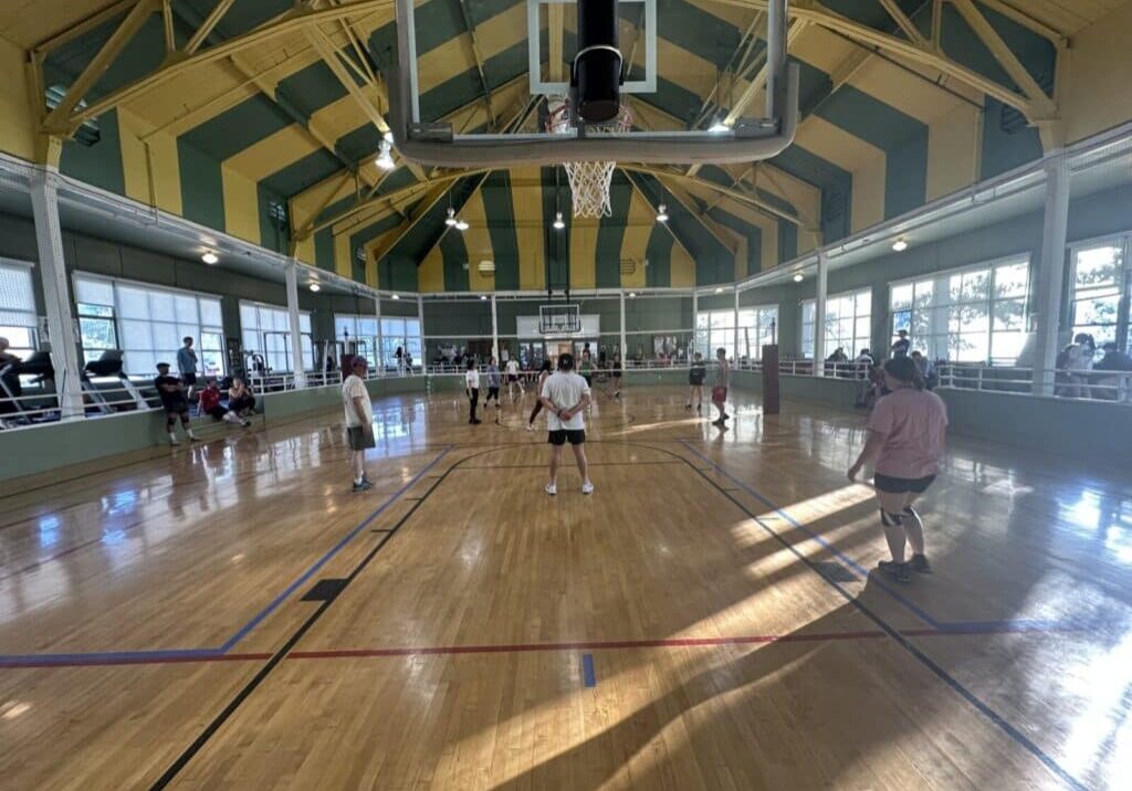 People play volleyball in a brightly lit indoor gym with a high ceiling, wooden floor, and a basketball hoop overhead. Spectators are seated along the sides.