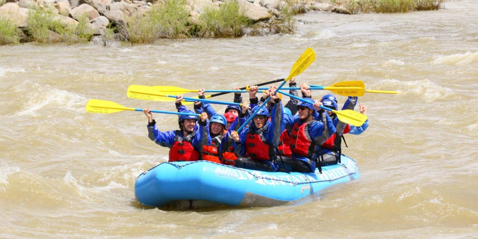 A group of people, including a Yellowstone employee, wearing helmets and life jackets holds paddles up while sitting in a blue inflatable raft on a river, ready to enjoy outdoor recreation together.