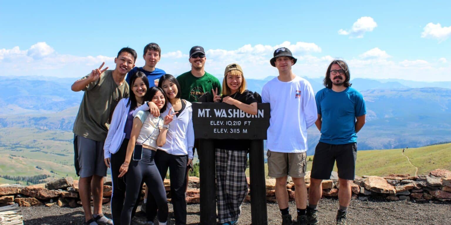 A group of eight people pose and smile around a Mt. Washburn summit sign in a mountainous outdoor setting under a blue sky.