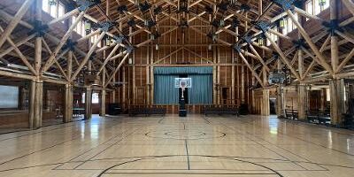 Indoor basketball court with wooden floor and exposed timber beams at the Old Faithful employee recreation center in Yellowstone, featuring a hoop in front of a stage and high windows letting in natural light.