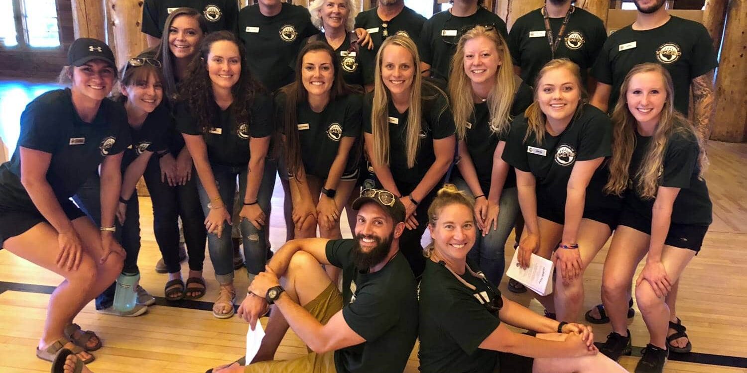 A group of 18 people wearing matching dark green shirts pose together indoors on a wooden floor, smiling at the camera.