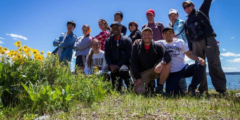A group of people from the Yellowstone employee recreation program pose together outdoors on a grassy area with yellow flowers, under a blue sky near a body of water.