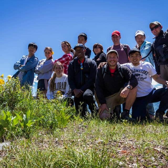 A group of people from the Yellowstone employee recreation program pose together outdoors on a grassy area with yellow flowers, under a blue sky near a body of water.
