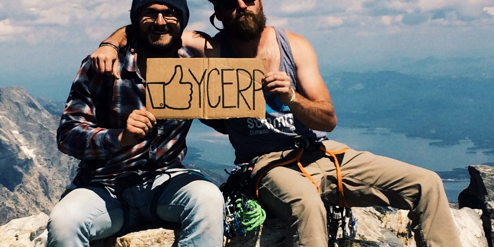 Two men, likely Yellowstone employees, sit atop a mountaintop in climbing gear, holding a "YCREP" thumbs-up sign. The scene captures outdoor recreation with a stunning mountain landscape in the background.