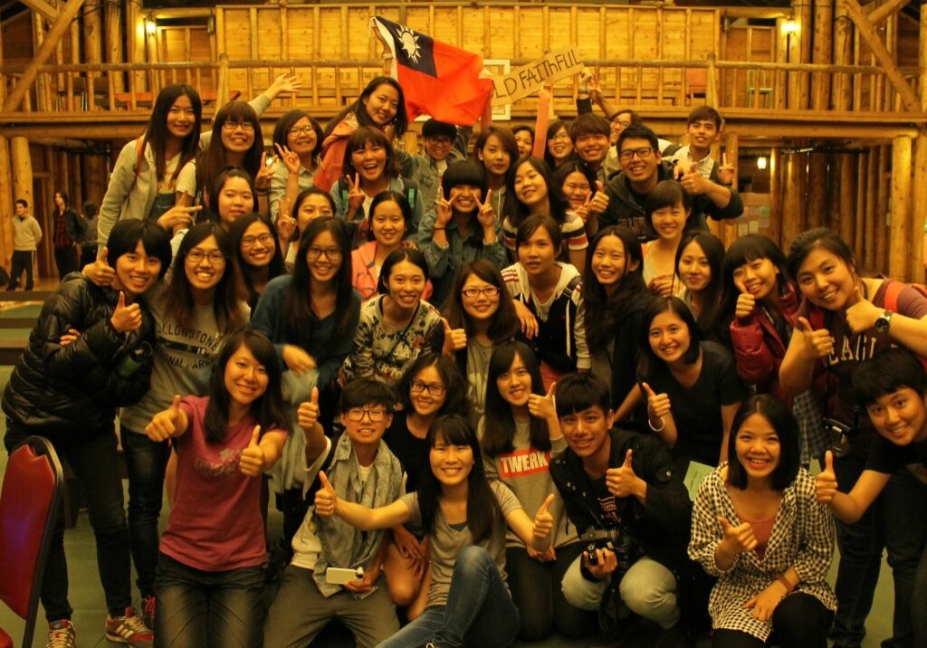 A large group of young people poses indoors, many giving thumbs up, with one person in the back holding a Taiwanese flag—capturing the spirit of the Yellowstone employee recreation program.