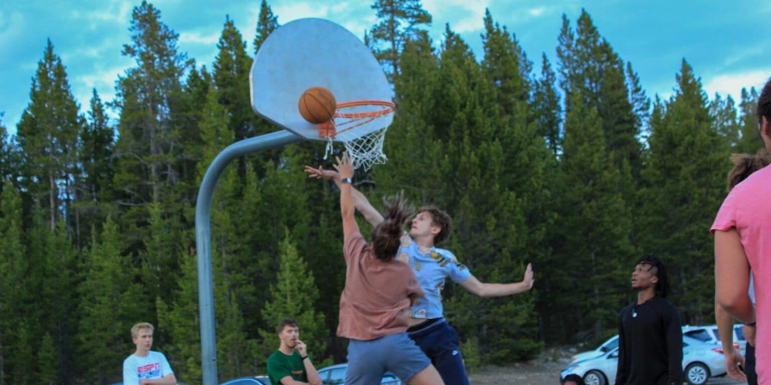 Two people jump for a basketball near an outdoor hoop, surrounded by trees and parked cars, as others—possibly Yellowstone employee athletics participants—watch the lively game.