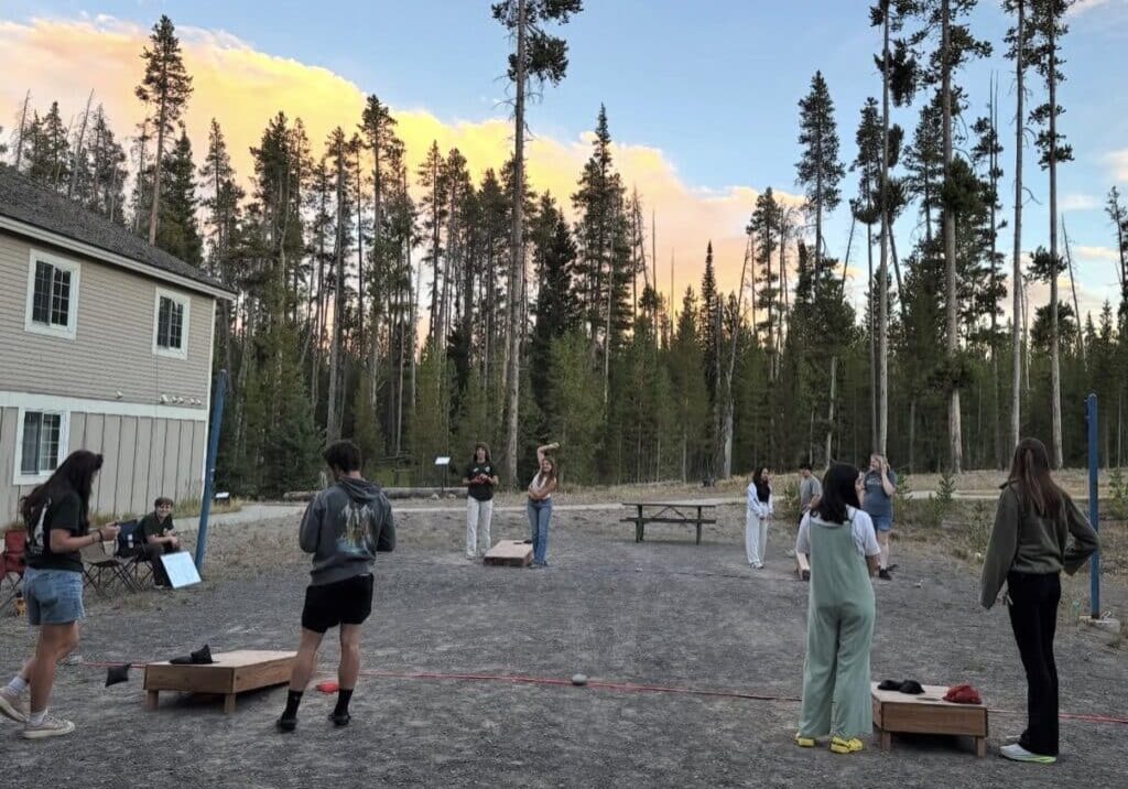 A group of people play cornhole and socialize outdoors near a building, surrounded by tall pine trees at sunset.