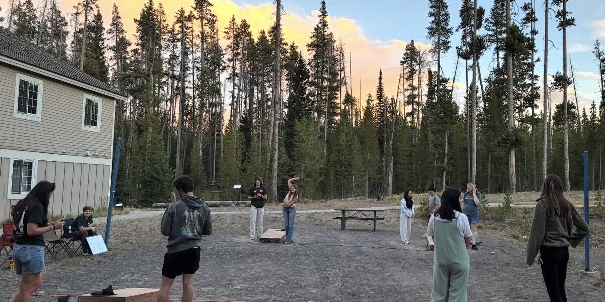 A group of people play cornhole and socialize outdoors near a building, surrounded by tall pine trees at sunset.