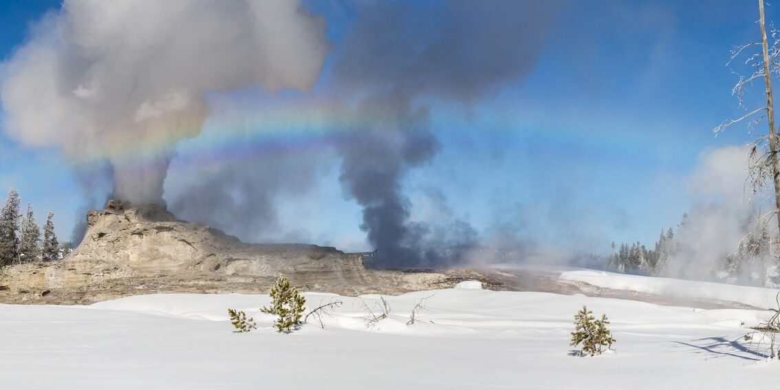 A geyser emits steam in a snowy landscape, with a rainbow visible in the mist and a clear blue sky above—an inspiring scene from the Yellowstone employee photo contest.