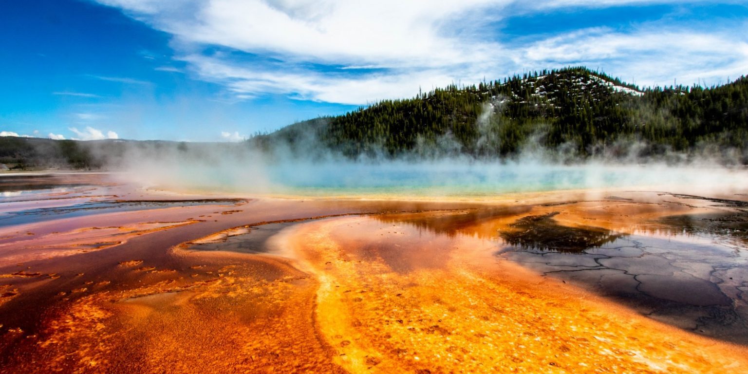 Colorful geothermal hot spring with orange mineral deposits, rising steam, and a forested hill under a partly cloudy sky.