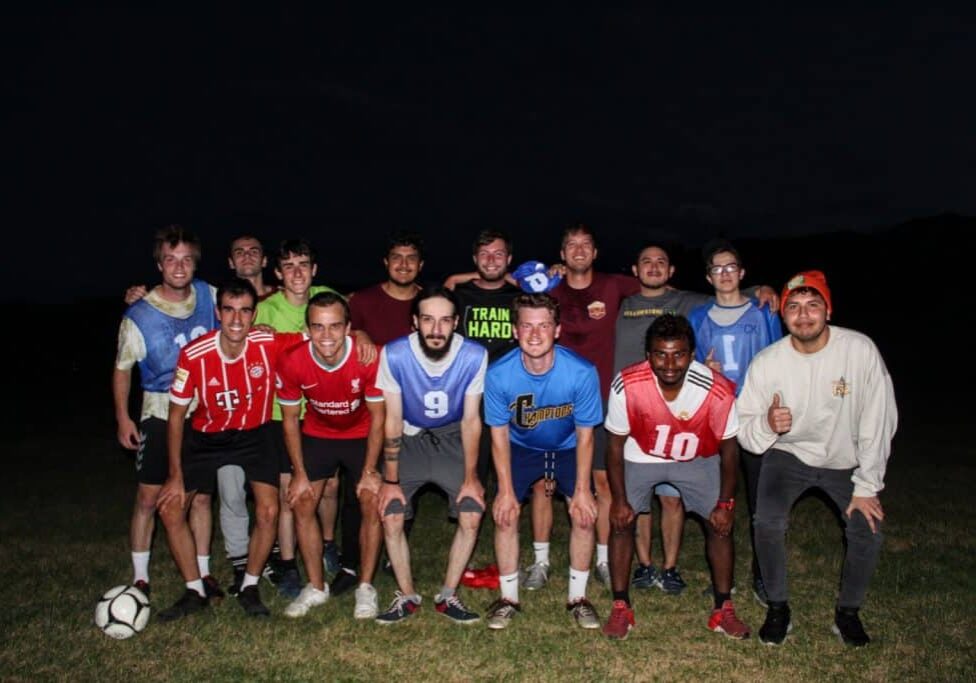 A group of sixteen men, part of Yellowstone employee athletics, pose together on a grassy field at night—some in soccer jerseys, others in casual clothes—with a soccer ball resting on the ground.